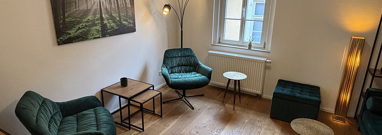 Modernes Sprechzimmer mit türkisfarbenen Sesseln, zwei Tischen und großem Fenster in heller, minimalistischer Einrichtung.